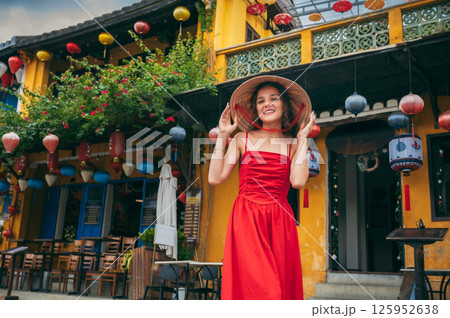 happy beautiful girl tourist in Vietnamese hat and red dress on the street in old town in Hoi An city in Vietnam in Asia 125952638