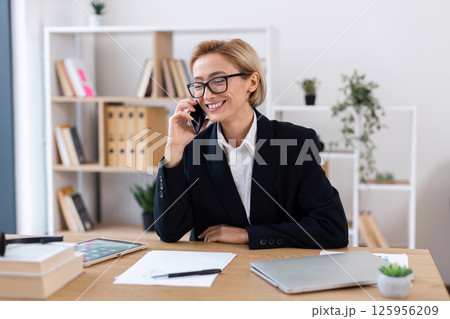 Professional woman in mid-thirties wearing business attire talking on phone at desk during work. Professional woman in mid-thirties wearing business attire talking on phone at desk during work. 125956209