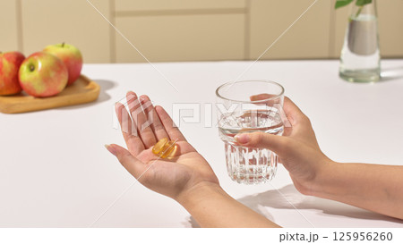 Capsule-shaped pills filled with nutrients are placed in the patient's palm, while the other hand holds a glass of water to take the medicine. Apples are on the white table. Healthcare concept. 125956260