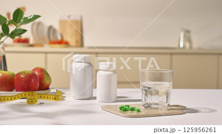 A glass of purified water sits on a tray with green pills. On the table are unlabeled pill bottles along with yellow. Body measuring tape and fresh apples. The concept of medicine in the kitchen. A glass of purified water sits on a tray with green pills. On the table are unlabeled pill bottles along with yellow. Body measuring tape and fresh apples. The concept of medicine in the kitchen. 125956261