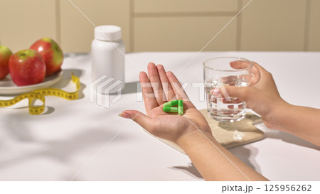 Weight loss pills come in capsule form and have a green shell, helping to improve body shape, placed in the hand, the other hand holds a glass of water to prepare to take the medicine. Weight loss pills come in capsule form and have a green shell, helping to improve body shape, placed in the hand, the other hand holds a glass of water to prepare to take the medicine. 125956262