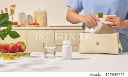 A woman in a blue shirt is taking a pill bottle out of bag. On the dining table are an unlabeled prescription bottle with tight-fitting lid, a glass of water, a plate of apples and a measuring tape. 125956263