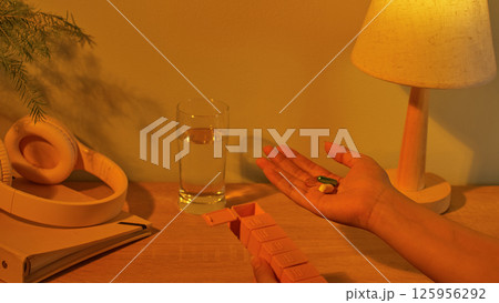 A daily pillbox with the first compartment open. The person in the photo holds two pills, ready to take before bed. Headphones are placed on a diary. The lighting in the room is a soft yellow. 125956292