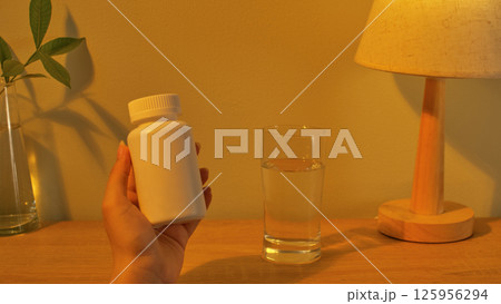 Mockup of a drug bottle being held by a hand, with the blank side facing the camera. Next to is a glass of water for easy medication. On the table is a lamp and a potted plant for decoration. 125956294