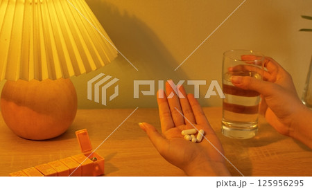A scene of taking medicine before bedtime. White pills in hand with a glass of water. The medicine helps patient sleep better. Night light with a spherical body, a pleated lampshade made of fabric. 125956295