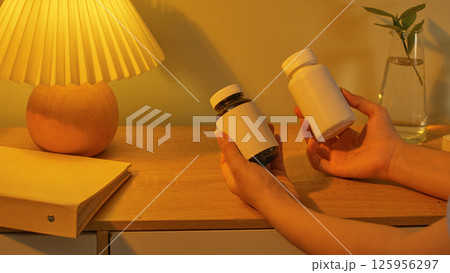 Unlabeled drug bottles in hand. A vintage draped-shaded nightlight is the only source of light illuminating the scene. A notebook and a potted plant are placed on the table. Warmly concept Unlabeled drug bottles in hand. A vintage draped-shaded nightlight is the only source of light illuminating the scene. A notebook and a potted plant are placed on the table. Warmly concept 125956297