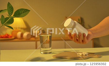 Photo of preparing to take dietary supplements in a cozy bedroom setting with pills prepared on a tray and a glass of purified water on the table. The light in the room is a warm and pale yellow. 125956302