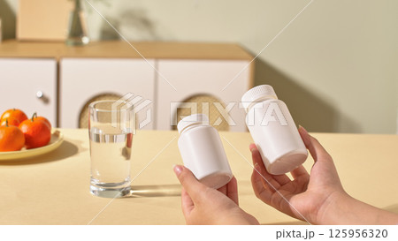 A person is holding two different sized medicine bottles. A glass of water is prepared on the table. A plate of fresh oranges with much vitamin C. The kitchen cabinets are blurred in the background. 125956320