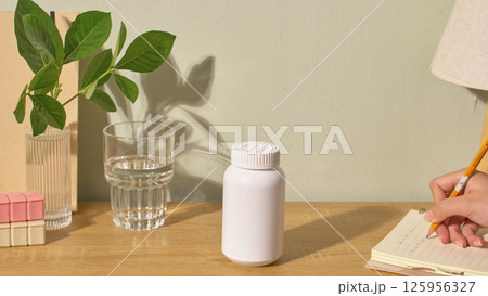 Mockup of a white drug bottle, used for advertising medical products. On the desk there is a glass of water, a vase, a branch and a notebook. The student is using a pencil to write on the notebook.  125956327