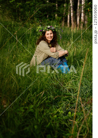 Woman in casual clothes and flower crown sitting in the grass, smiling and holding berry. Rural eco-lifestyle, summer joy and unplugged simplicity 125959060