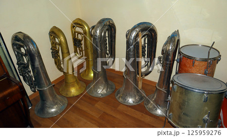 Old brass instruments, including tubas and a drum, are neatly arranged in a music school storage room, showcasing the history of musical education. Old brass instruments, including tubas and a drum, are neatly arranged in a music school storage room, showcasing the history of musical education. 125959276