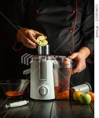 A skilled chef is preparing a fresh juice by placing slices of apple into a juicer. The warm kitchen is filled with fruits, creating a cozy atmosphere during the evening A skilled chef is preparing a fresh juice by placing slices of apple into a juicer. The warm kitchen is filled with fruits, creating a cozy atmosphere during the evening 125959651