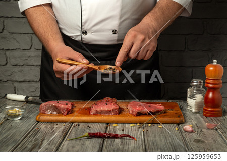 A chef prepares raw meat by carefully sprinkling seasoning from a spoon, showcasing culinary skills. The kitchen is well-lit, enhancing the focus on the fresh ingredients 125959653