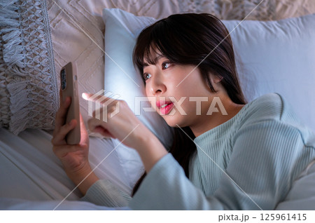 布団の中で横になりながらスマホを見て指を動かす女性の様子 125961415