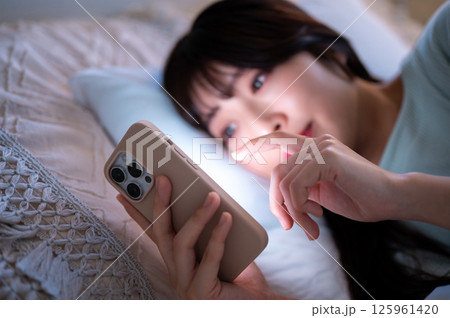 暗い寝室でスマートフォンの画面を指でスクロールする女性の様子 125961420
