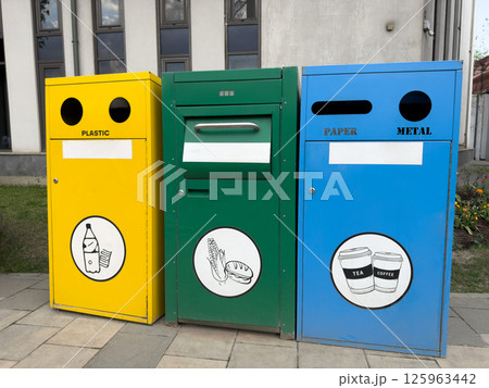 Three colorful recycling bins for plastic, organic waste, and paper or metal stand on pavement near modern building, promoting environmental awareness, sustainable habits, and urban waste separation Three colorful recycling bins for plastic, organic waste, and paper or metal stand on pavement near modern building, promoting environmental awareness, sustainable habits, and urban waste separation 125963442