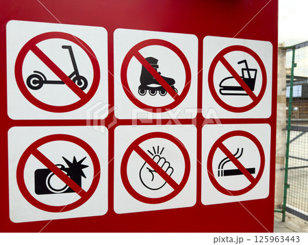 Closeup of red prohibition signboard with six warning icons indicating no scooters, rollerblades, bumper cars, flash photography, loud noises, or smoking in restricted area near entrance 125963443