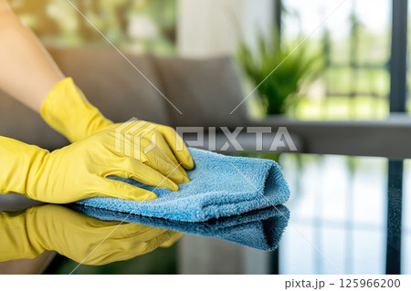 person hands in yellow rubber gloves carefully wipe glass table with blue microfiber cloth in bright living room 125966200