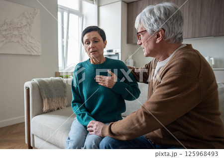 Wife health issues. Mature woman grab chest struggles for air comforted on couch by supportive man Wife health issues. Mature woman grab chest struggles for air comforted on couch by supportive man 125968493