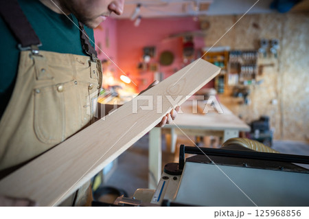 Concentrated attentive craftsman checking wooden plank carefully before crafting in carpentry 125968856