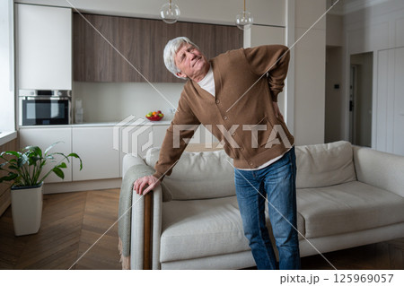 Struggling anguished elderly pensioner grimaces in pain from backache renal colic, leaning on sofa Struggling anguished elderly pensioner grimaces in pain from backache renal colic, leaning on sofa 125969057
