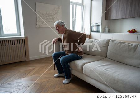 Lonely unhealthy elderly man holds aching back unable to rise from couch due to spinal problems Lonely unhealthy elderly man holds aching back unable to rise from couch due to spinal problems 125969347