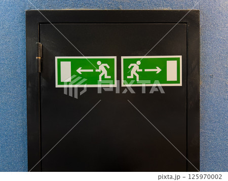 Two emergency exit signs with running figure and arrows pointing left and right placed on black door panel against blue wall, providing clear direction for safe evacuation in case of emergency Two emergency exit signs with running figure and arrows pointing left and right placed on black door panel against blue wall, providing clear direction for safe evacuation in case of emergency 125970002