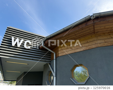 Modern public restroom building with large WC letters on metal facade and wooden roof detail under blue sky, combining practical function with clean architectural design in outdoor urban space Modern public restroom building with large WC letters on metal facade and wooden roof detail under blue sky, combining practical function with clean architectural design in outdoor urban space 125970016