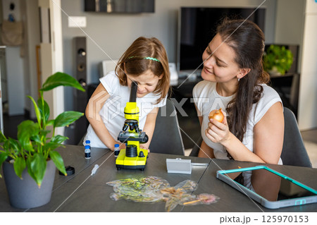 Little girl and her mother examining onion skin under a microscope at home. Concept of family science education and early biology learning Little girl and her mother examining onion skin under a microscope at home. Concept of family science education and early biology learning 125970153