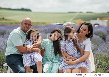 A family sits on a bench in the middle of a lavender field. The two twins are wearing identical purple dresses, the older sister is wearing a green dress in a lavender field. A family sits on a bench in the middle of a lavender field. The two twins are wearing identical purple dresses, the older sister is wearing a green dress in a lavender field. 125970950