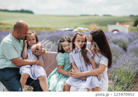 A family sits on a bench in the middle of a lavender field. The two twins are wearing identical purple dresses, the older sister is wearing a green dress in a lavender field. A family sits on a bench in the middle of a lavender field. The two twins are wearing identical purple dresses, the older sister is wearing a green dress in a lavender field. 125970952