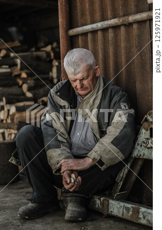 A pensioner rests on the ground, his gaze lost in thought. The elderly man wears clothes that have seen many years of work. 125971921