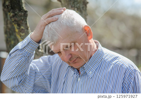 A portrait of a pensioner stroking his gray hair. A sad old man standing by a tree. 125971927