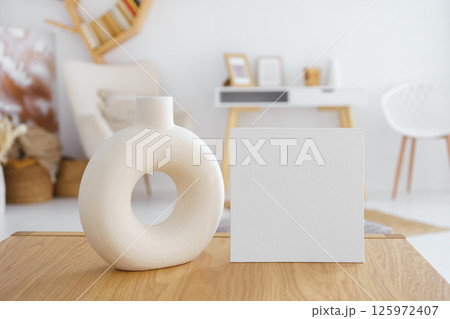 Decorative vase and blank white cardboard on wooden table in bright living room. Mockup, template 125972407