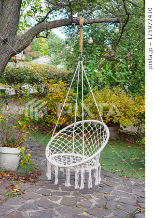 Comfortable hanging wicker white chair, hammock chair in boho style hung on a tree in summer garden Comfortable hanging wicker white chair, hammock chair in boho style hung on a tree in summer garden 125972410