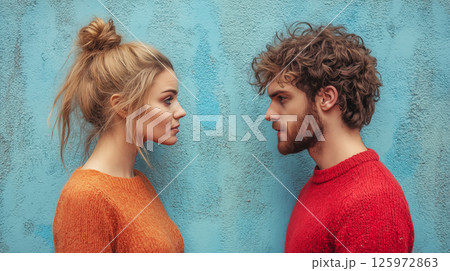 Young couple facing each other against blue wall wearing orange and red sweaters with contrasting hairstyles. Concept of relationship, communication, and emotional connection 125972863