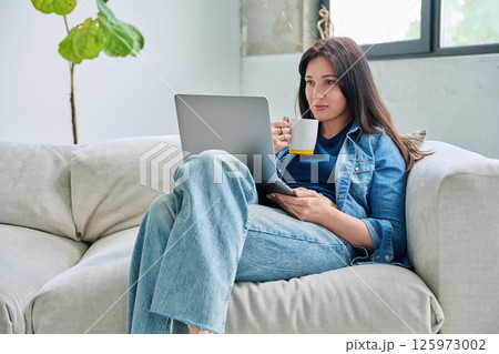 Young relaxed woman sitting on sofa at home with cup, looking at laptop 125973002