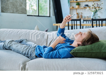 Serious teenager guy looking at smartphone screen lying on the couch at home Serious teenager guy looking at smartphone screen lying on the couch at home 125973021