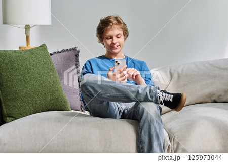 Handsome smiling teenage guy using smartphone sitting on couch at home Handsome smiling teenage guy using smartphone sitting on couch at home 125973044