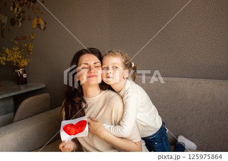 Young girl with light hair hugs mom tightly while holding up her childish drawing. Pure mother-daughter love. 125975864