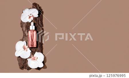 Glass cosmetic bottle on textured tree bark podium with white orchid flowers in rays of sunlight with hard shadows. 125976995
