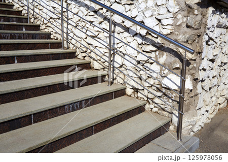 A solid staircase made of grey concrete and decorated with metal railings stands against a rugged stone wall. 125978056