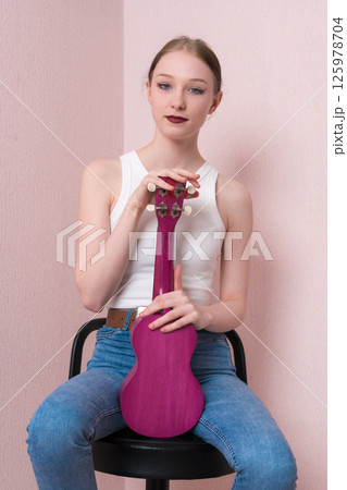 Portrait of calm young female musician with ukulele held upright, sitting on bar stool and looking confidently at camera. Beautiful woman in casual clothing with white singlet and blue jeans Portrait of calm young female musician with ukulele held upright, sitting on bar stool and looking confidently at camera. Beautiful woman in casual clothing with white singlet and blue jeans 125978704