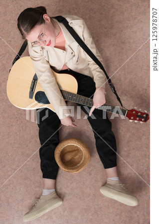 High angle view of guitarist with guitar, sitting with spread legs and straw hat in front of her, intended as donation for playing music. Woman looking camera, feeling of hope, pleasant anticipation 125978707