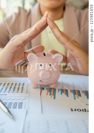 Close-up of hands forming a roof over a pink piggy bank on financial documents with graphs, symbolizing savings and financial security. 125981959