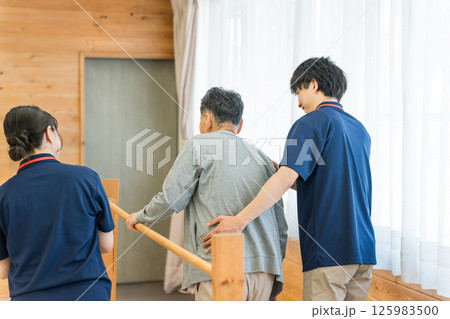手すりを使い歩行訓練するシニア男性と介助する作業療法士,理学療法士の男性スタッフ 125983500