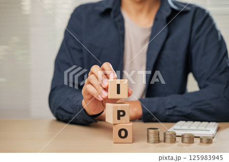 Close up businessman hand holding wooden cube blocks spelling IPO near coins and a calculator. Represents Initial Public Offering, planning, employee initiatives, and business financial accountability Close up businessman hand holding wooden cube blocks spelling IPO near coins and a calculator. Represents Initial Public Offering, planning, employee initiatives, and business financial accountability 125983945