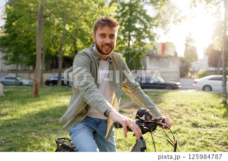 Young man enjoying a sunny day while biking in a park near urban surroundings and tall trees 125984787
