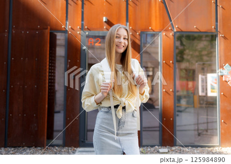 Young woman poses outside a modern building wearing a yellow blouse and denim skirt during daylight hours 125984890
