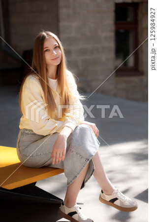 Young woman sitting on a yellow bench in sunlight wearing casual attire with a relaxed expression in an urban setting Young woman sitting on a yellow bench in sunlight wearing casual attire with a relaxed expression in an urban setting 125984927